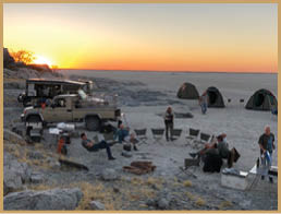 Two men are sitting in chairs under a tent in a sandy area. A truck is parked nearby. There are also two tents and a car in the scene. AI generated content