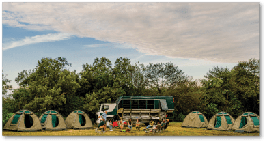 A group of tents are set up in a field, with a person standing in front of them. The tents are arranged in a circle, and there is a truck nearby. The sky is cloudy, and the scene appears to be set in a wooded area. AI generated content