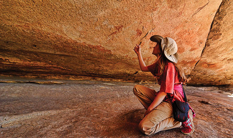 A woman is sitting on a rock, pointing at a cave. She is wearing a hat and a red shirt. AI generated content