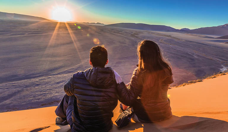 A man and woman sitting on a sandy hill. Description generated by AI