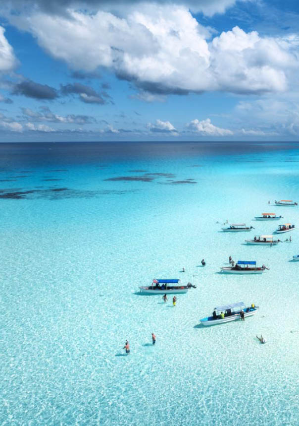 Aerial view of colorful boats in clear azure water in summer. Mnemba island, Zanzibar. Top drone view of sandbank in low tide, blue sea, white sand, swimming people, yachts, sky with clouds. Ocean